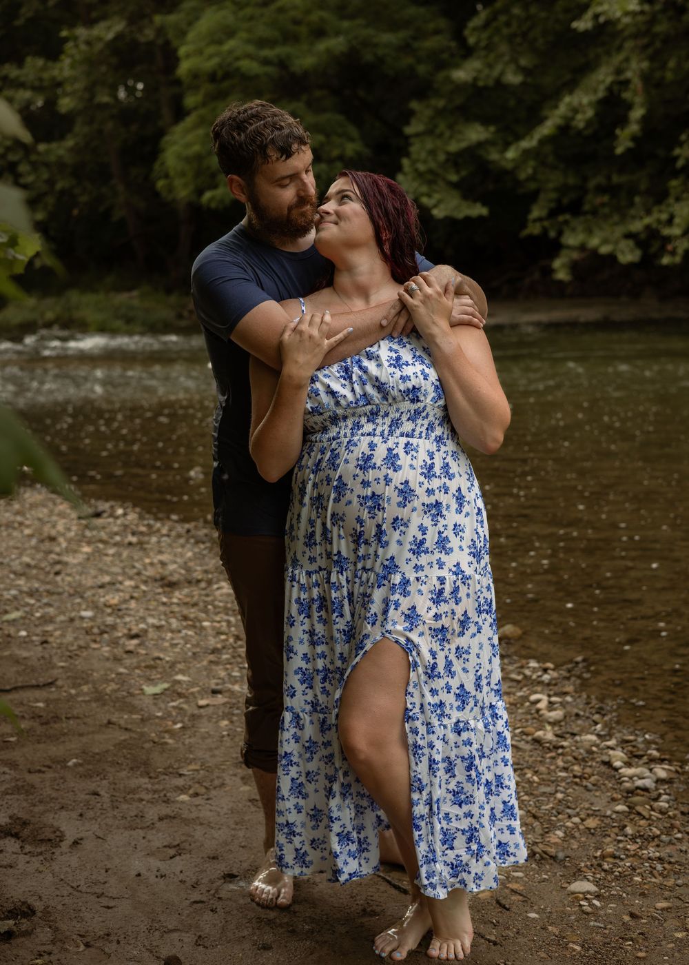 Bearded man kissing womans cheek from behind as she laughs joyfully near a forest river.