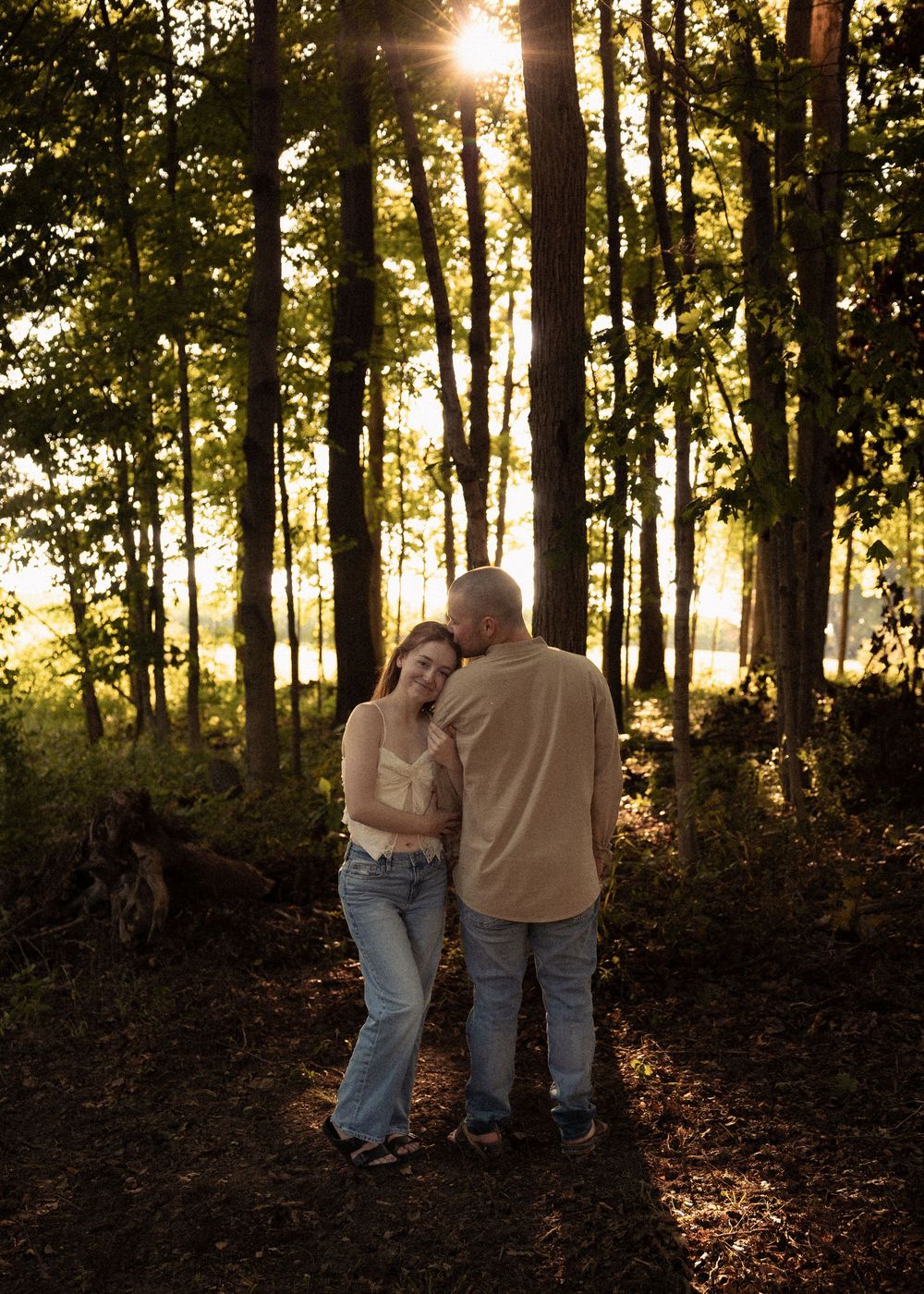Couple sharing a tender moment in a sunlit forest, golden light streaming through tall trees.