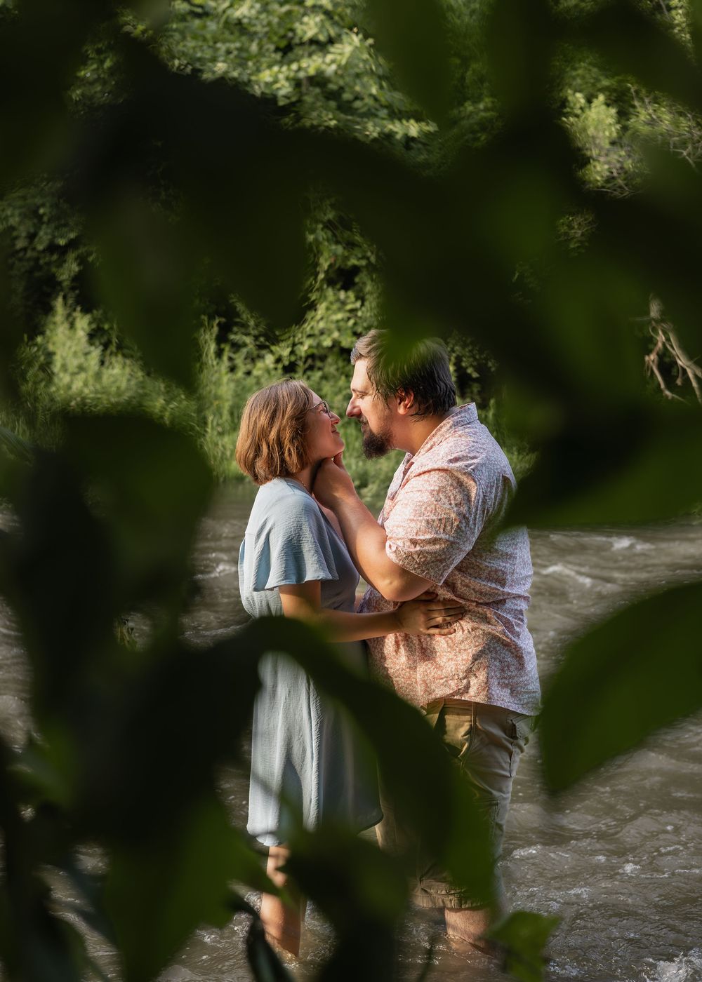 Romantic couple embracing near a stream, viewed through lush green leaves in soft natural light.