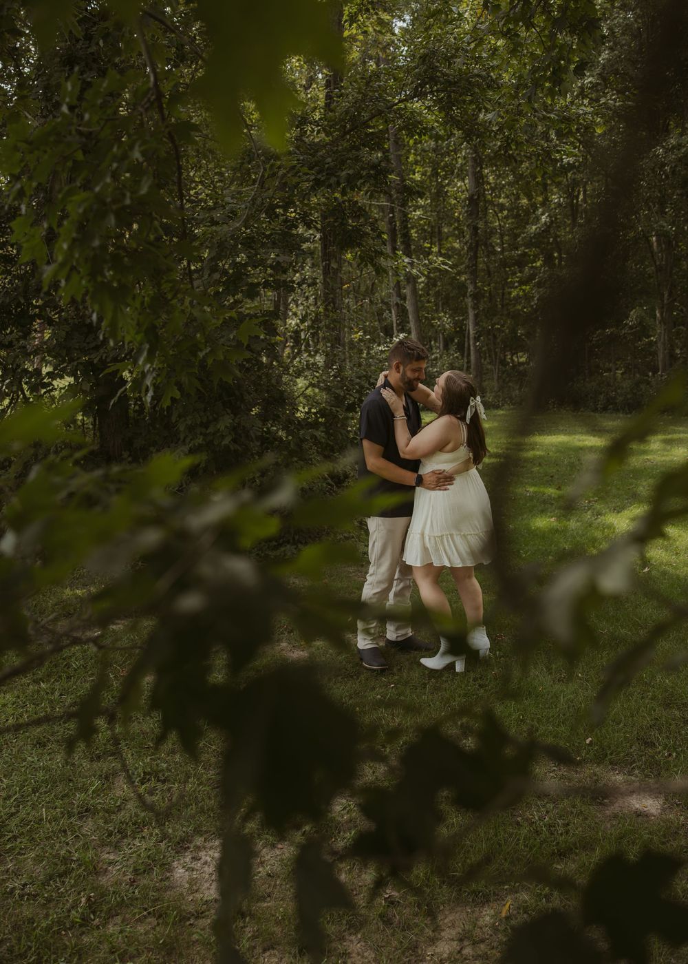 Couple sharing a romantic kiss in a lush green forest, framed by leaves, man in black shirt, woman in white dress.