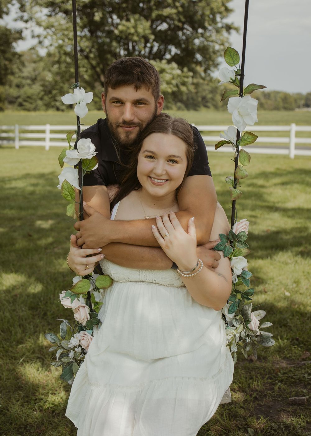 Couple posing on a floral swing outdoors, woman in white dress smiling, man embracing her from behind on green lawn.