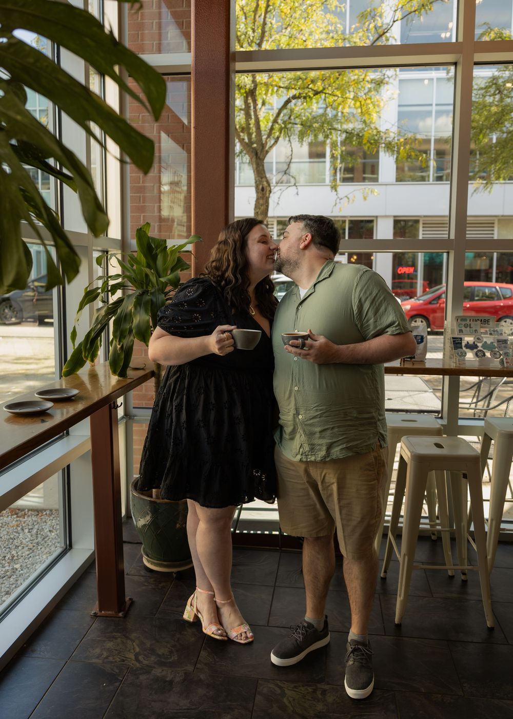 Couple sharing a kiss in a modern cafe, holding coffee cups near large windows with city views.