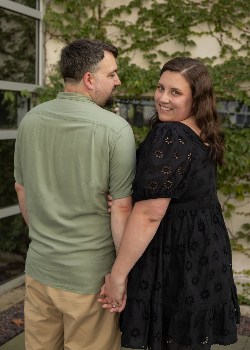 Couple holding hands outdoors, woman in black floral dress smiling, man in green polo and khaki pants, ivy-covered building.