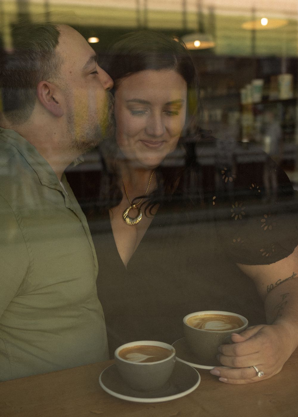 Couple sharing an intimate moment over coffee cups at a cafe table, photographed through a window with reflections.