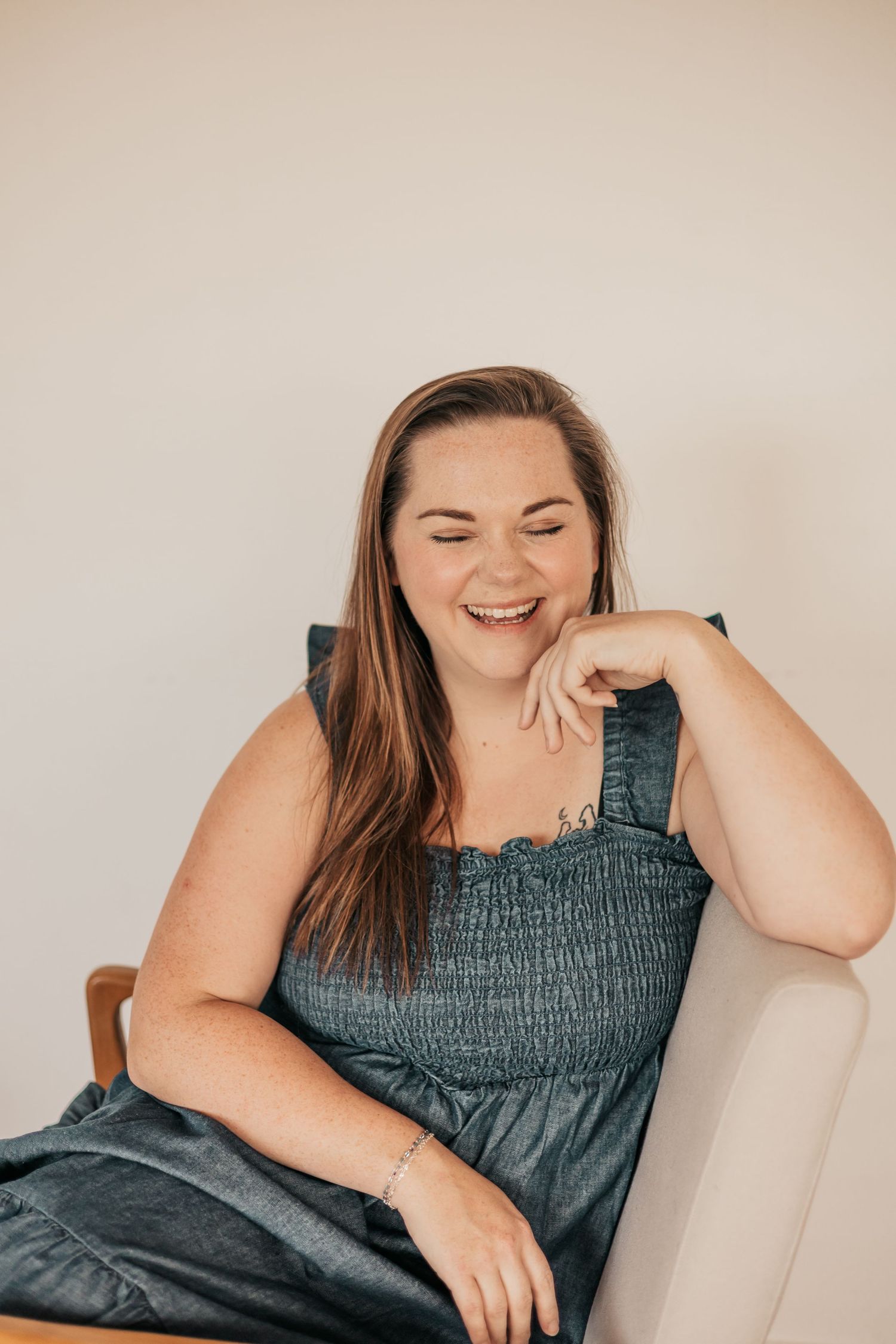 A smiling woman with long brown hair sits relaxed in a chair, wearing a teal smocked dress with ruffle details | Photography Business Coach