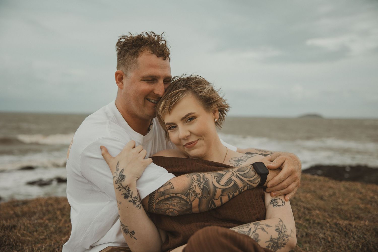 Couple with tattoos embracing on rocky beach with ocean waves and cloudy sky behind them.