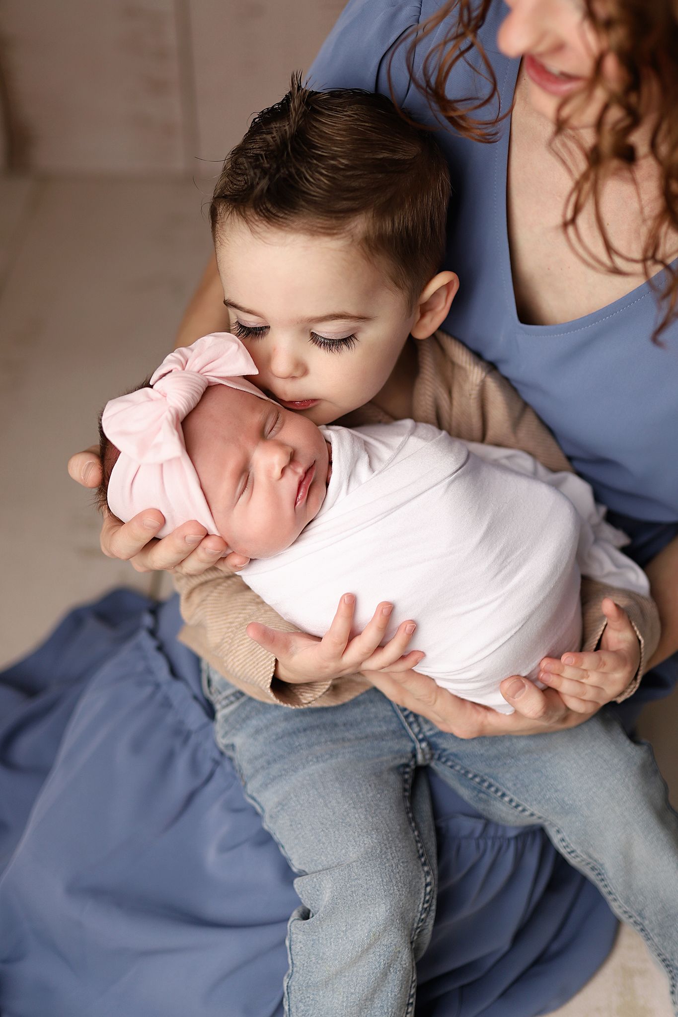 A toddler gently kisses a sleeping newborn wrapped in white, held by their mother in a blue dress.