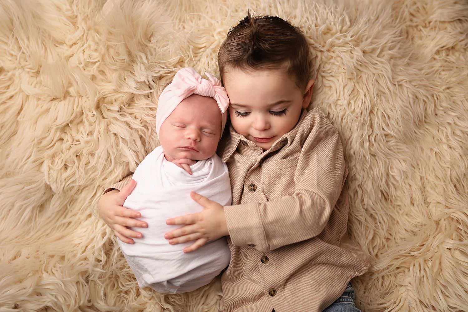 Toddler boy gently holding sleeping newborn baby girl in pink bow on fluffy cream fur backdrop.