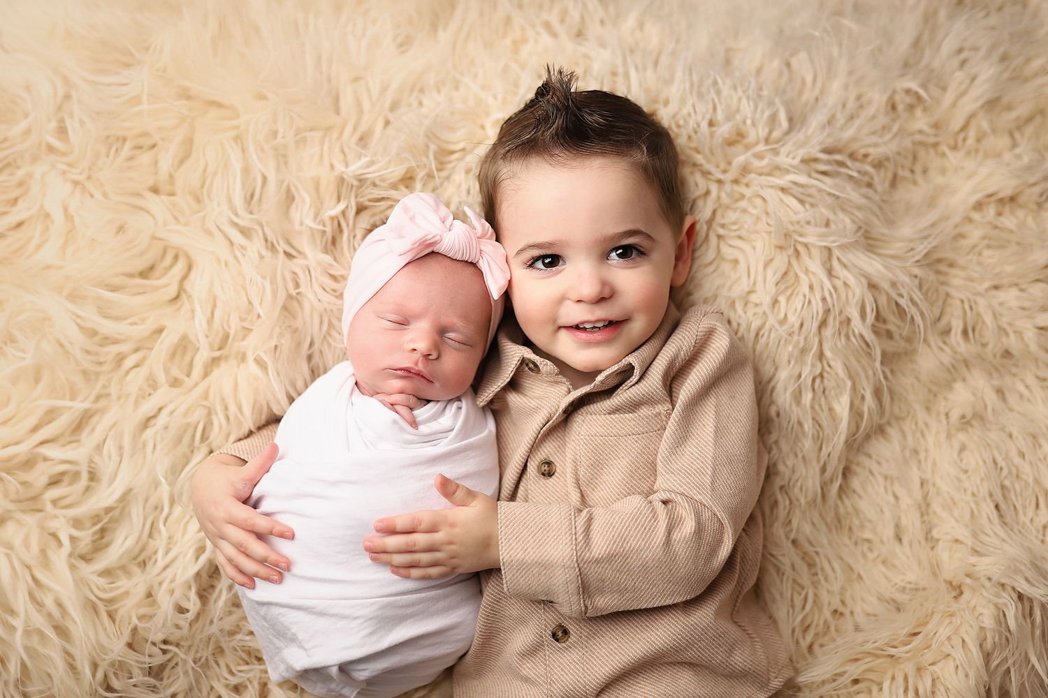 Toddler boy in beige shirt smiling while gently holding swaddled newborn baby girl with pink bow on fur backdrop. Orem newborn photographer