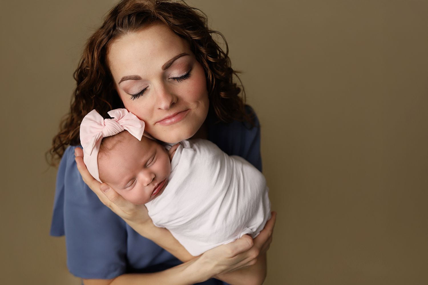 A mother with curly brown hair tenderly holds her swaddled newborn baby girl wearing a pink bow headband.