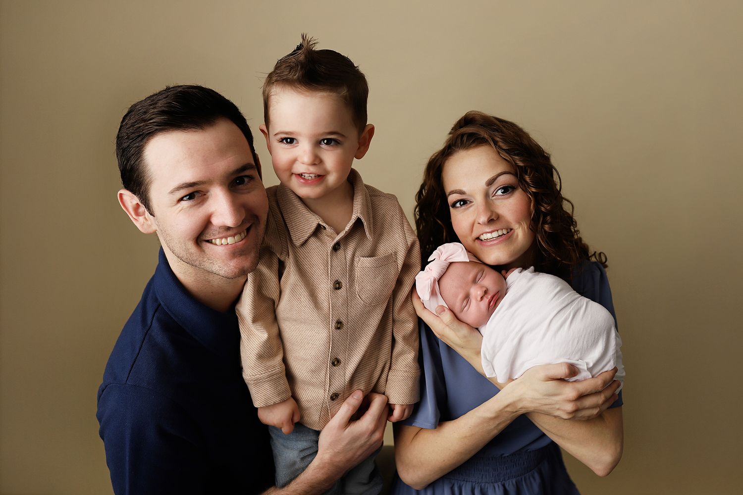 A smiling family of four poses for a studio portrait, with parents holding a toddler and a newborn baby. Orem newborn photography