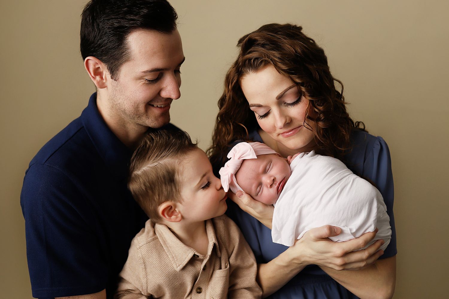Family portrait of parents with toddler son kissing newborn baby sister wrapped in white swaddle.