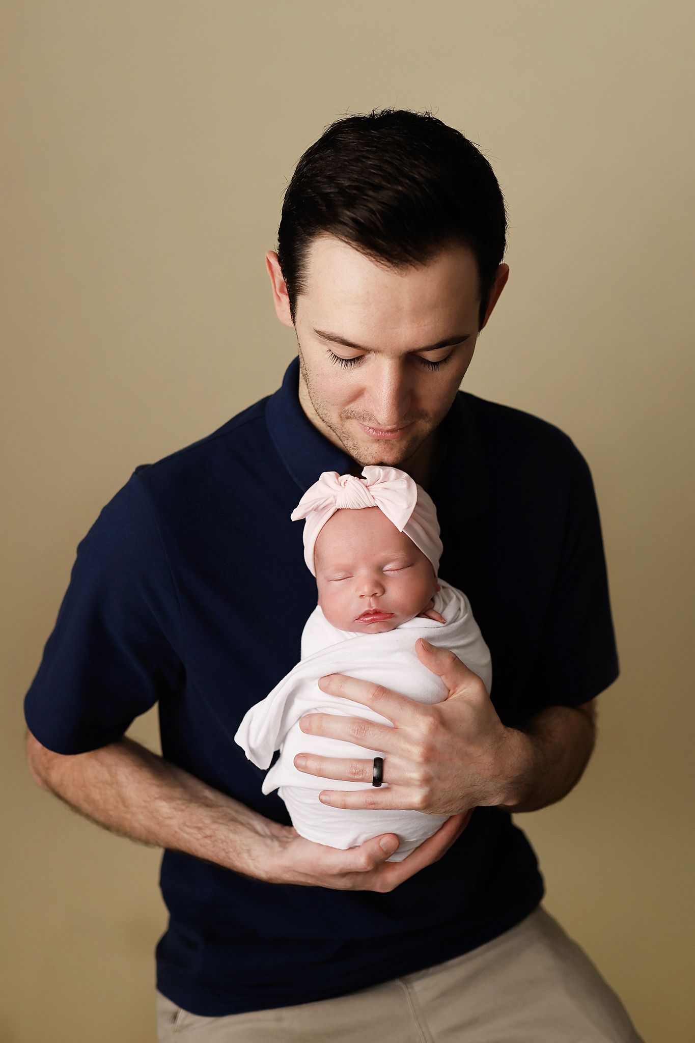 A father tenderly holds his swaddled newborn baby girl wearing a pink bow headband against a neutral background.