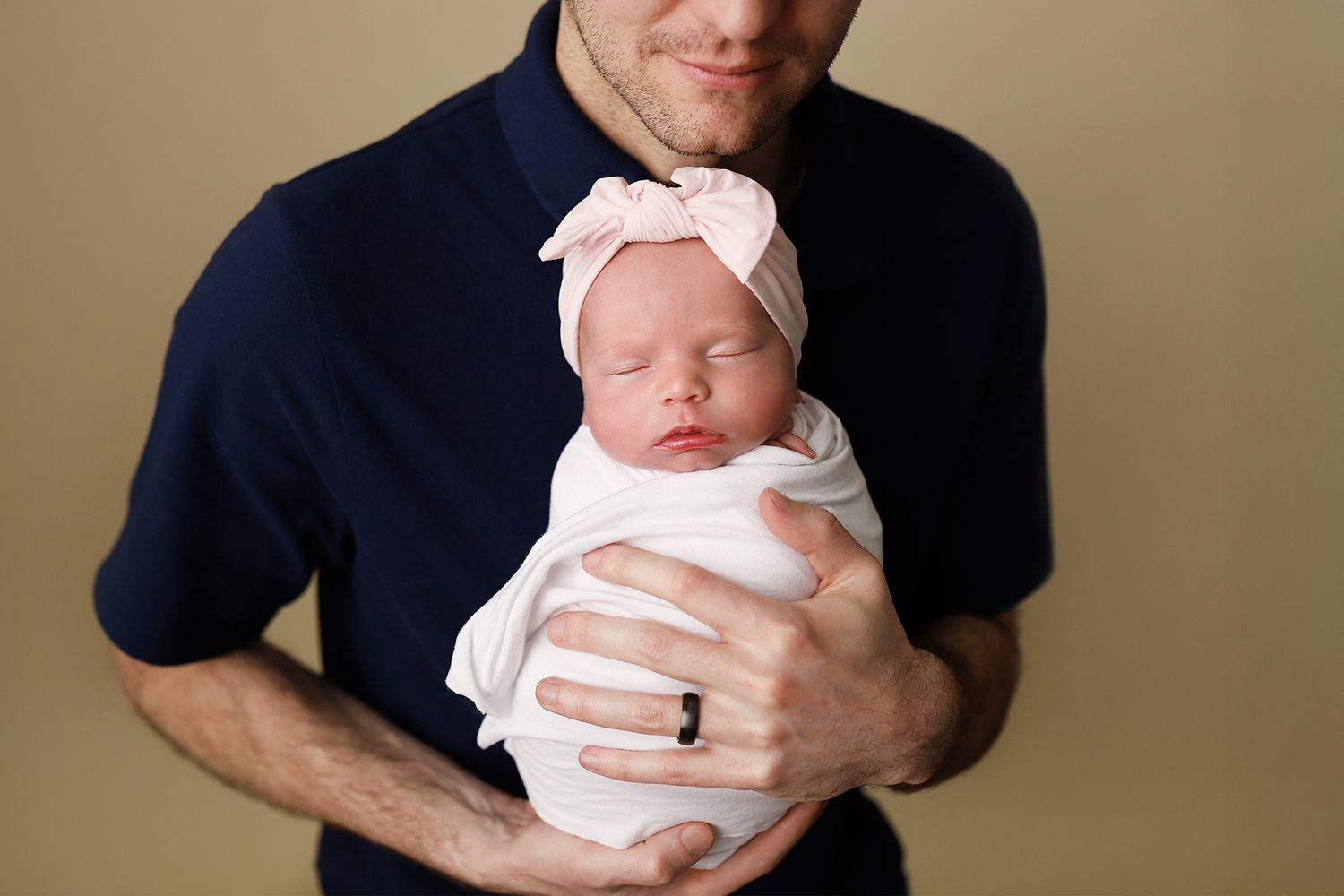 Father in navy shirt cradling swaddled newborn baby girl wearing pink bow headband against beige background.