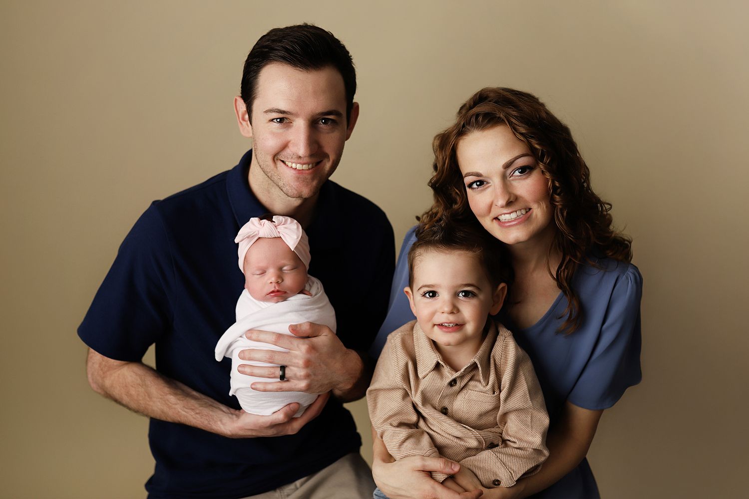 Happy family portrait with parents holding newborn baby and toddler son, smiling against neutral background.