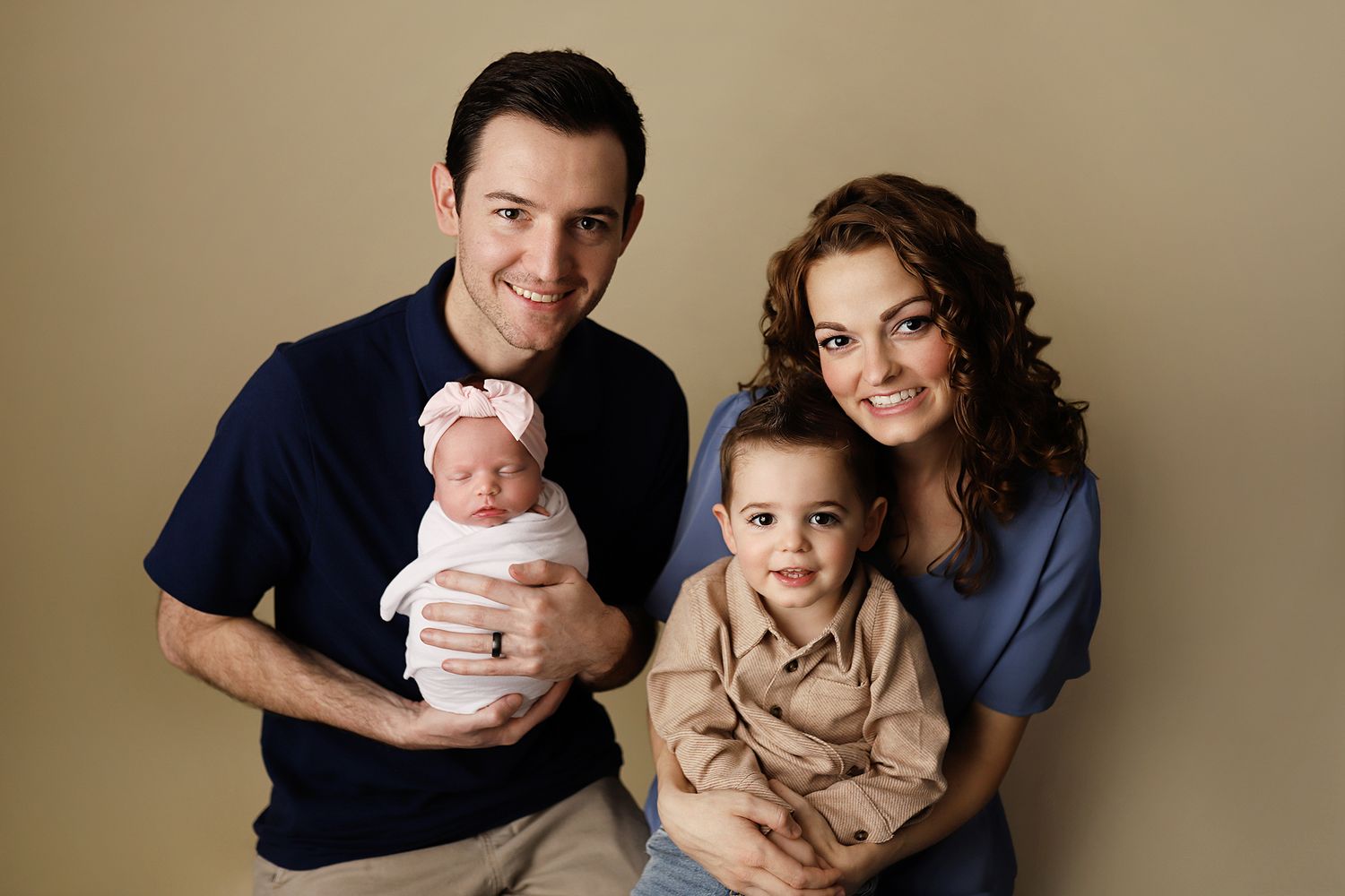 Happy family portrait with parents holding newborn baby and toddler son against neutral beige background. Orem newborn photographer