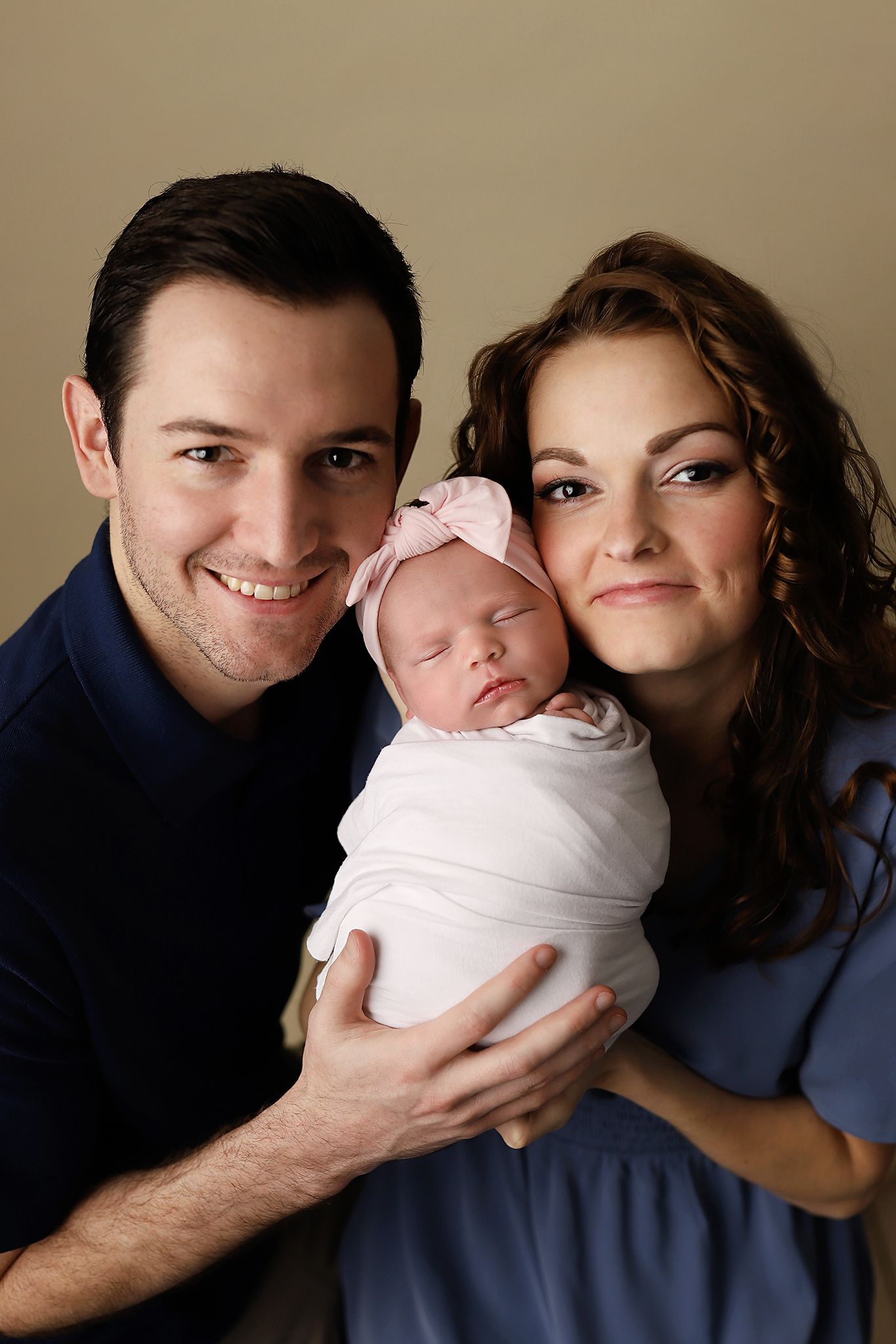 Happy parents smiling and holding their sleeping newborn baby girl wrapped in white with a pink bow headband.
