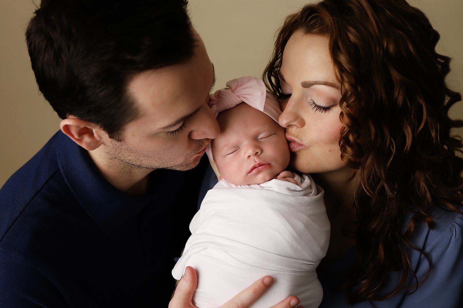 New parents kissing their sleeping newborn baby wrapped in white swaddle with pink bow headband. Newborn studio in Orem
