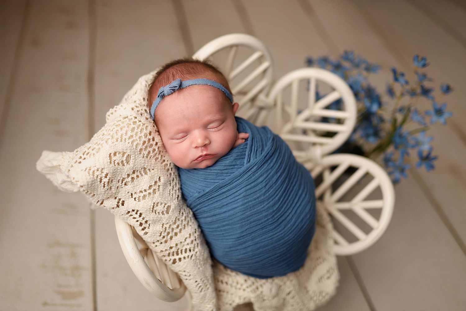 Newborn baby swaddled in blue wrap sleeping in a white wooden cart with lace blanket and blue flowers. Newborn photography studio in Orem