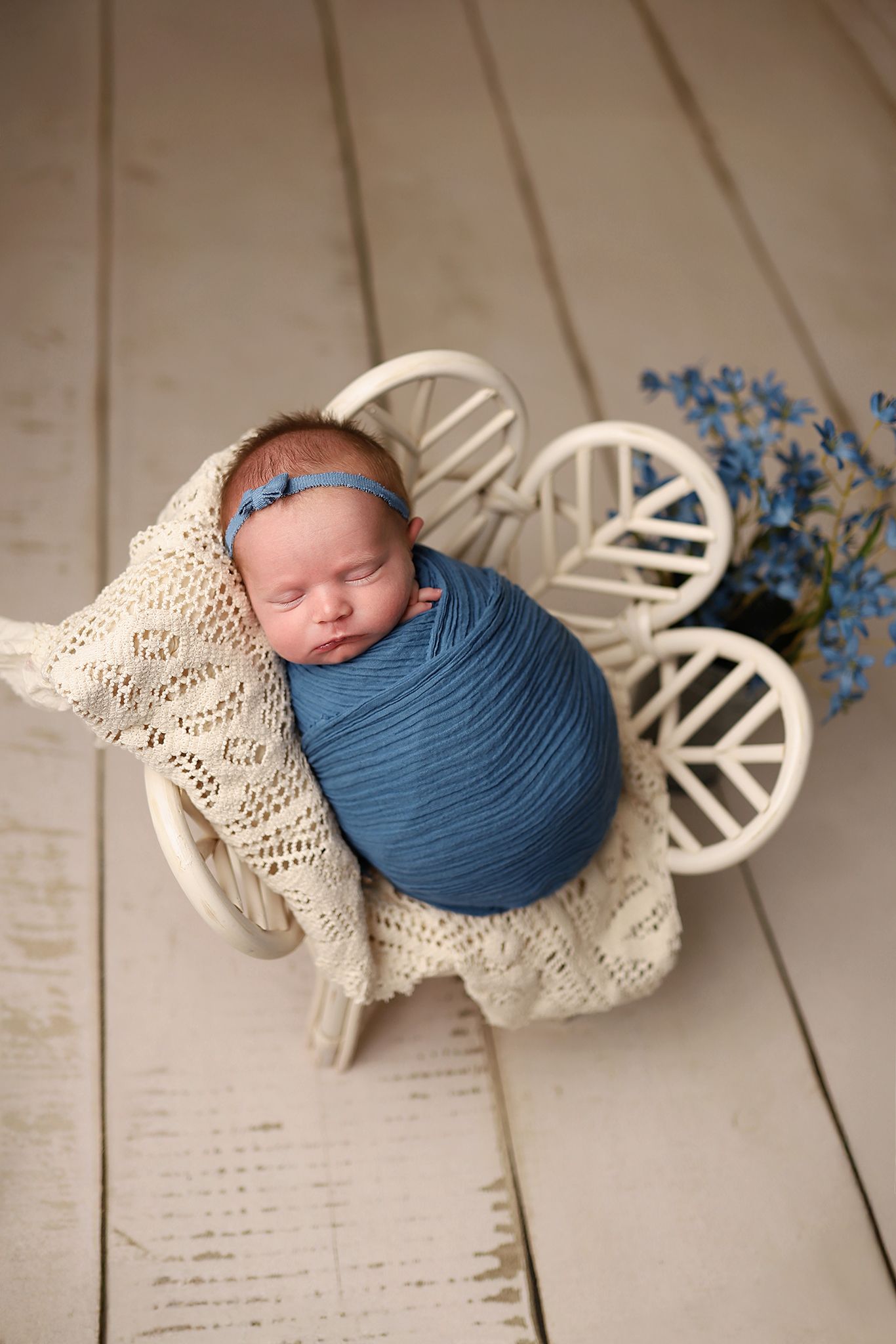 Sleeping newborn baby swaddled in blue wrap, resting in white wicker chair with cream knit blanket and blue flowers.