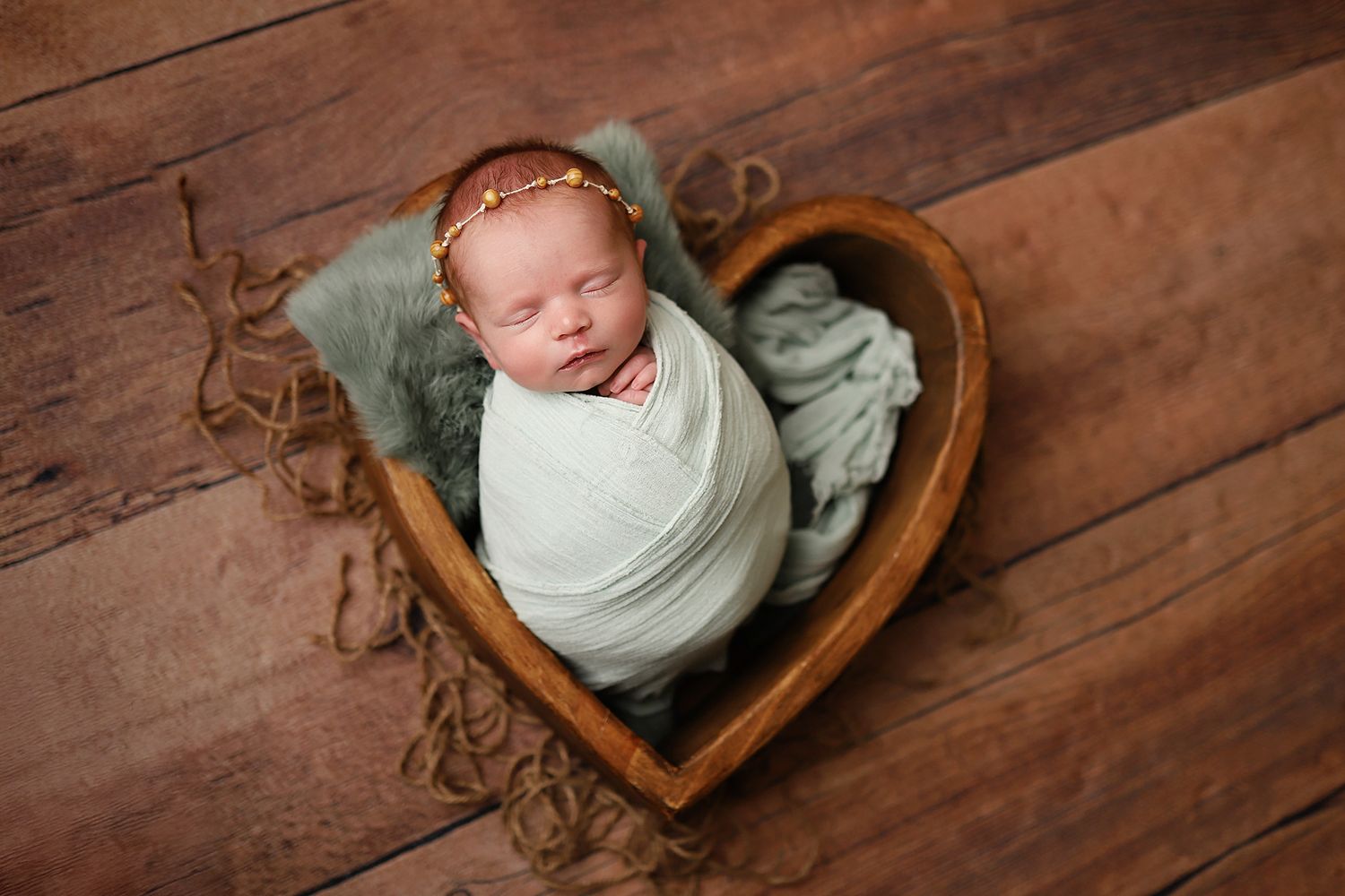 Newborn baby swaddled in white wrap sleeping in a wooden heart-shaped bowl with a floral headband. Orem newborn photography