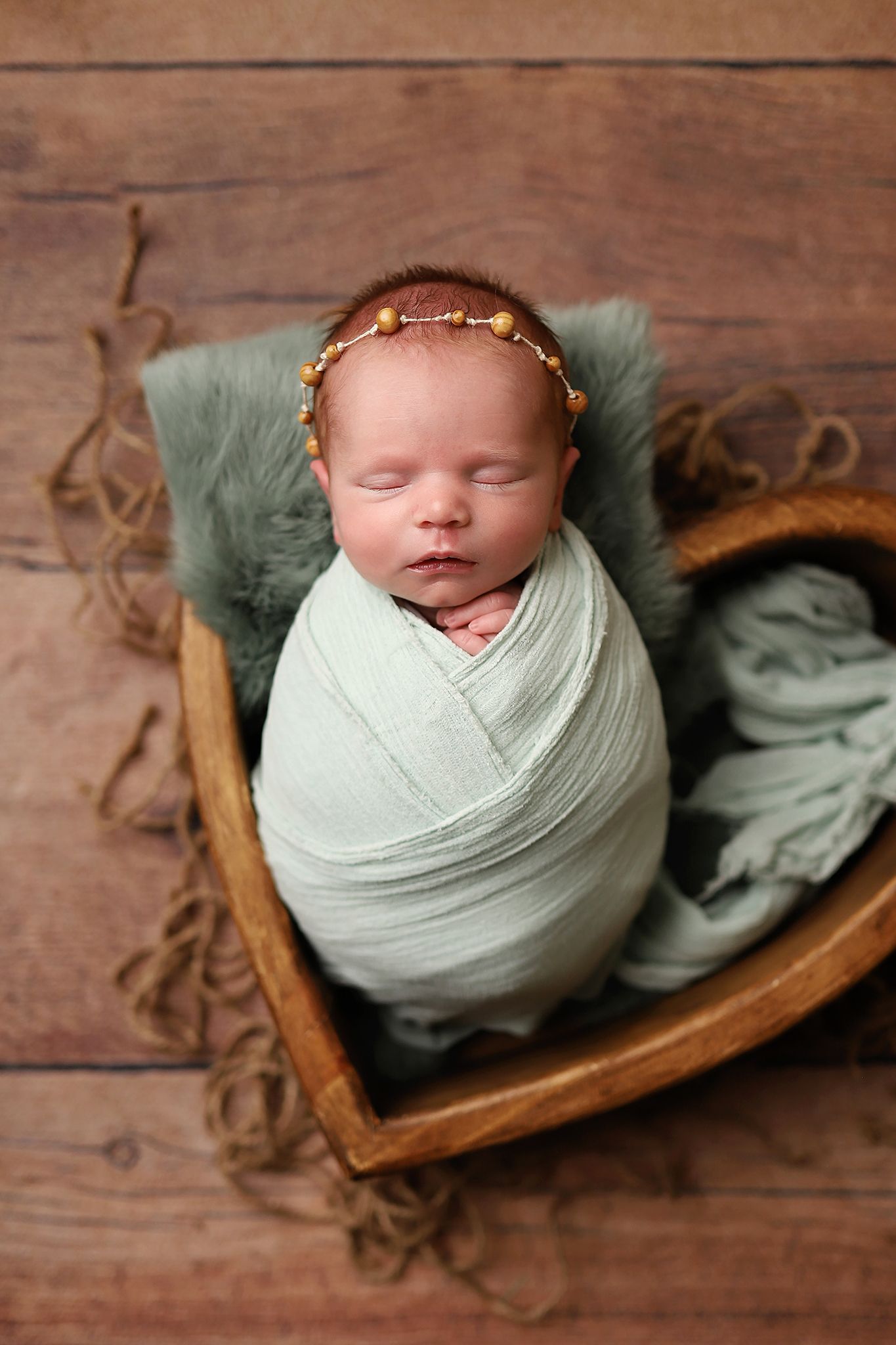 Newborn baby swaddled in mint green wrap, sleeping in heart-shaped wooden bowl with floral headband.