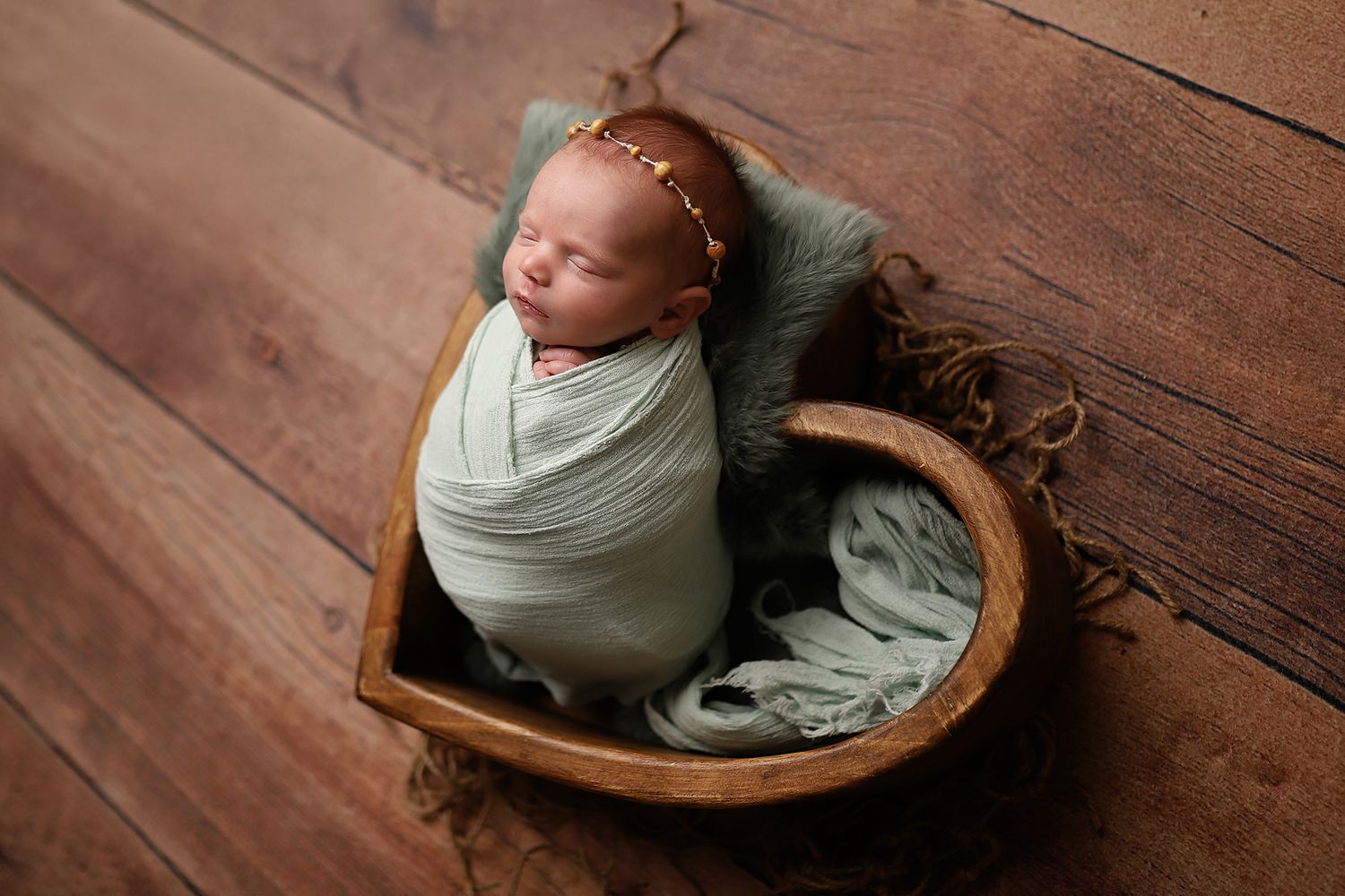 Newborn baby swaddled in sage green wrap, sleeping peacefully in a wooden heart-shaped bowl on hardwood floor.