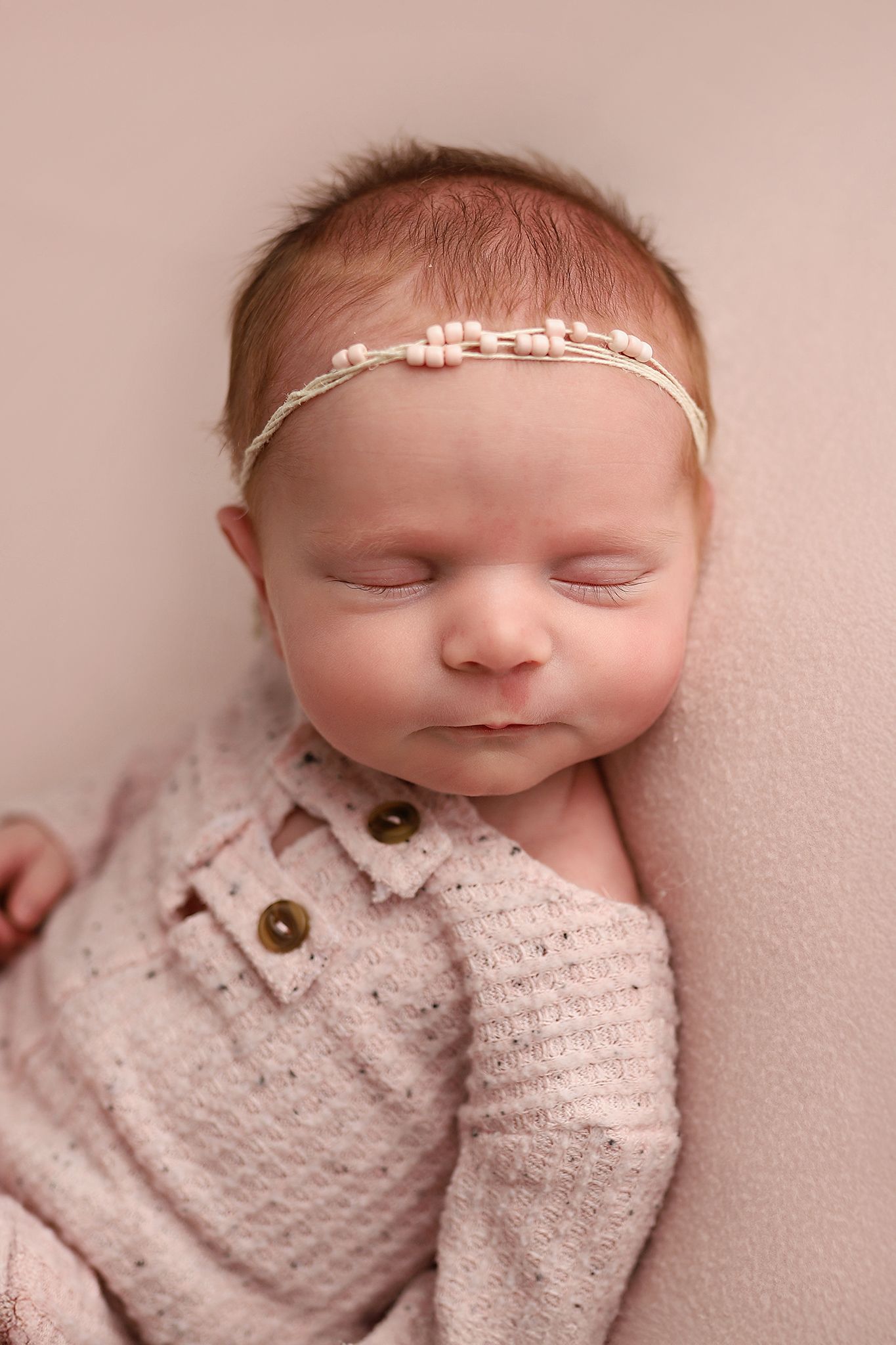 Sleeping newborn baby girl in pink knit romper and delicate headband on soft pink blanket.
