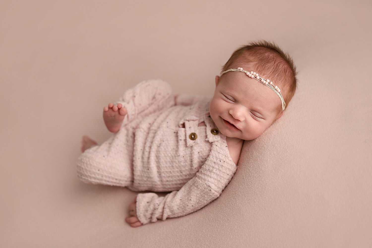 Smiling newborn baby girl in pink knit romper and floral headband lying on beige backdrop.