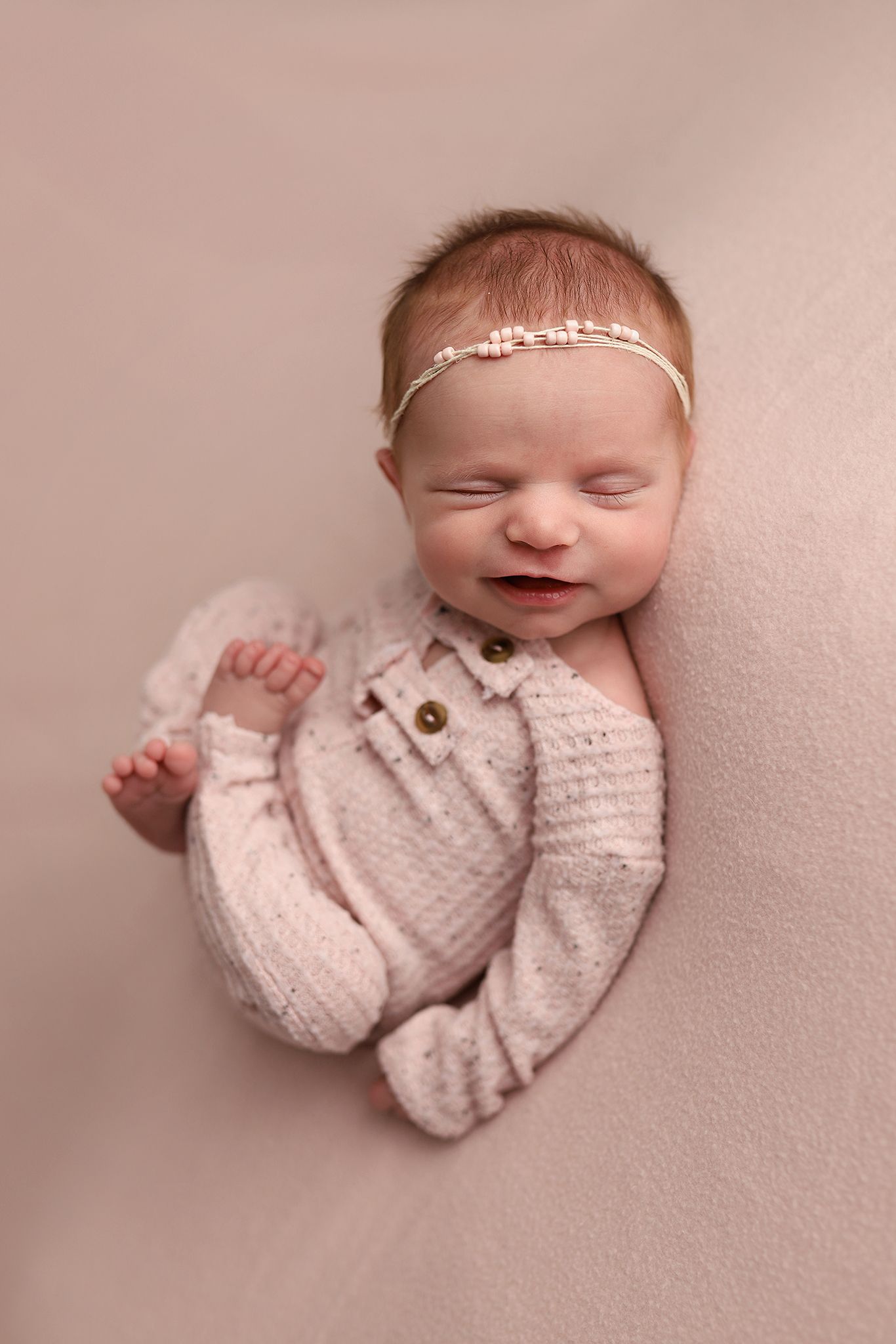 Smiling newborn baby girl in pink knit romper and floral headband sleeping peacefully on a soft beige backdrop. Orem newborn photographer