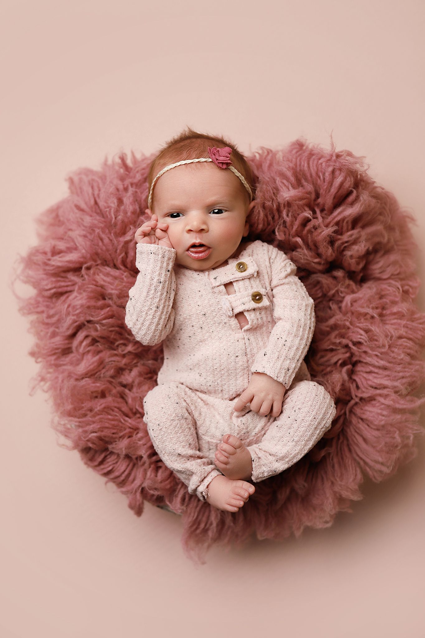 Newborn baby girl in pink knit romper and floral headband lying on dusty rose fluffy round rug.