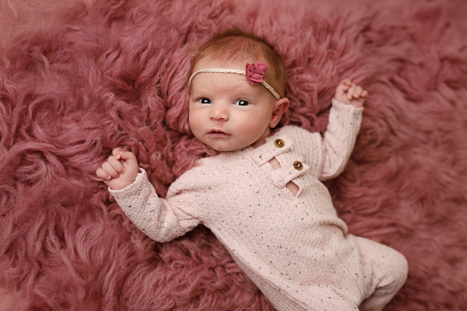 Adorable newborn baby girl in pink knit romper and floral headband lying on fluffy mauve fur rug.