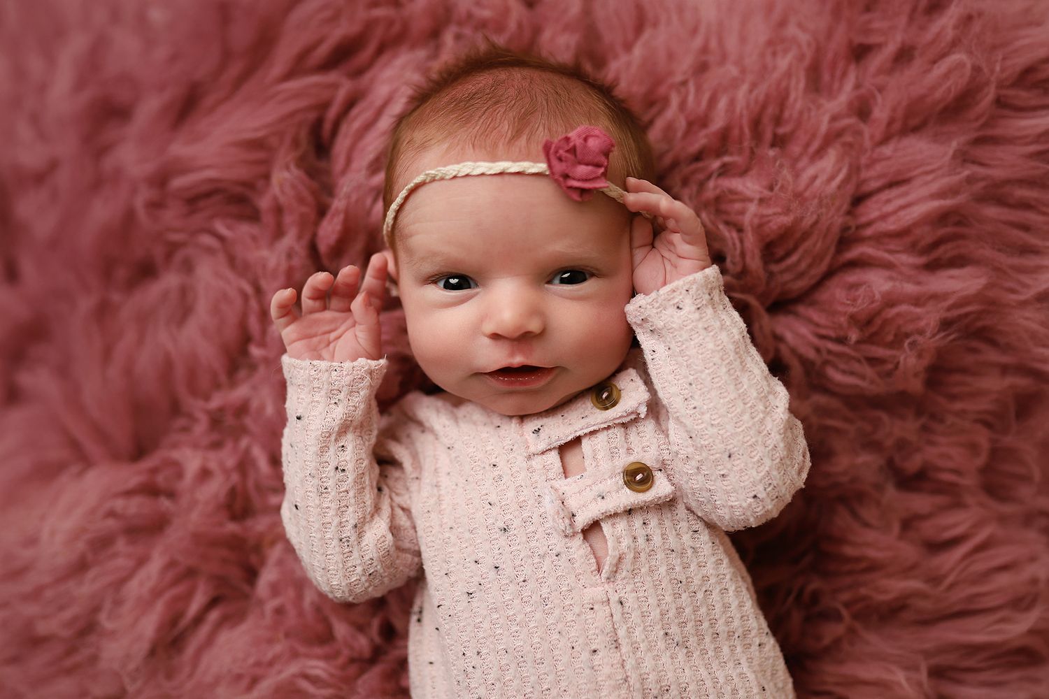Smiling newborn baby girl in pink knit outfit on fluffy mauve rug wearing pink floral headband. Orem newborn photographer
