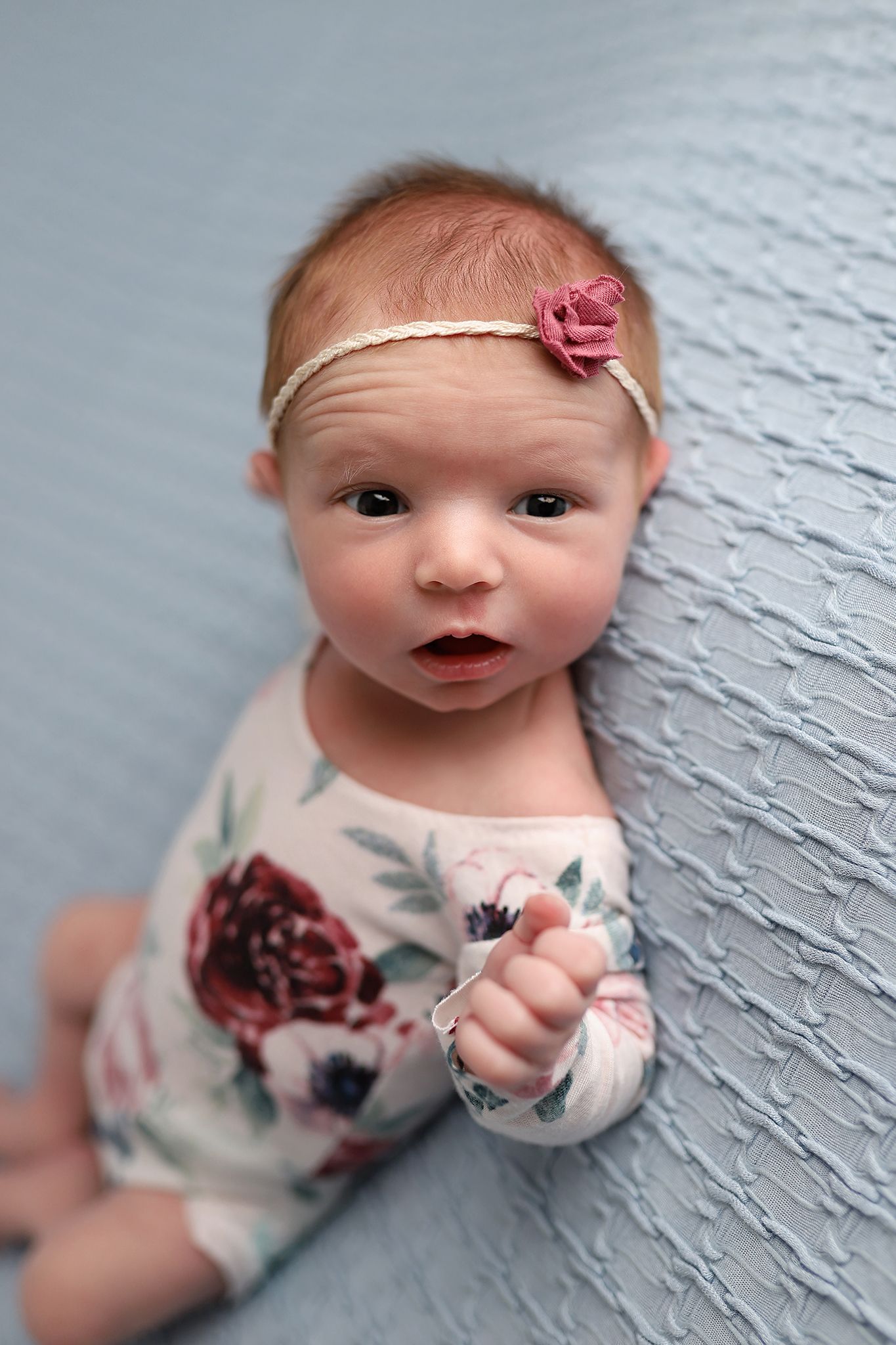 Newborn baby girl in floral outfit and pink flower headband lying on blue textured blanket.