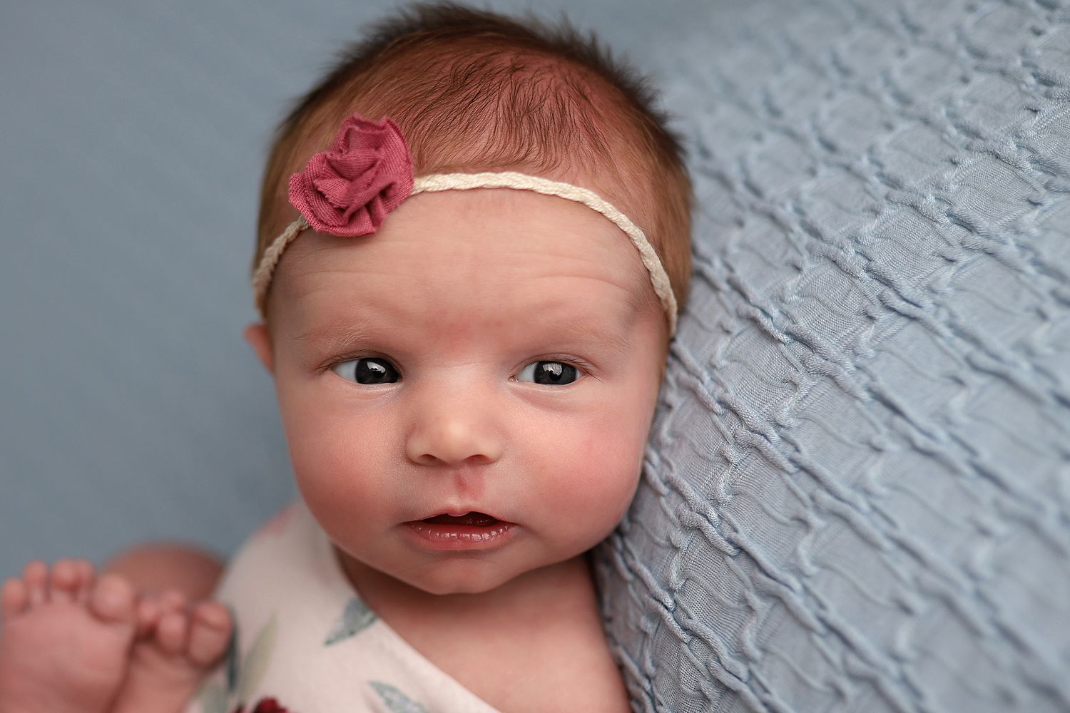 Newborn baby girl wearing a pink floral headband lying on a soft blue textured blanket.