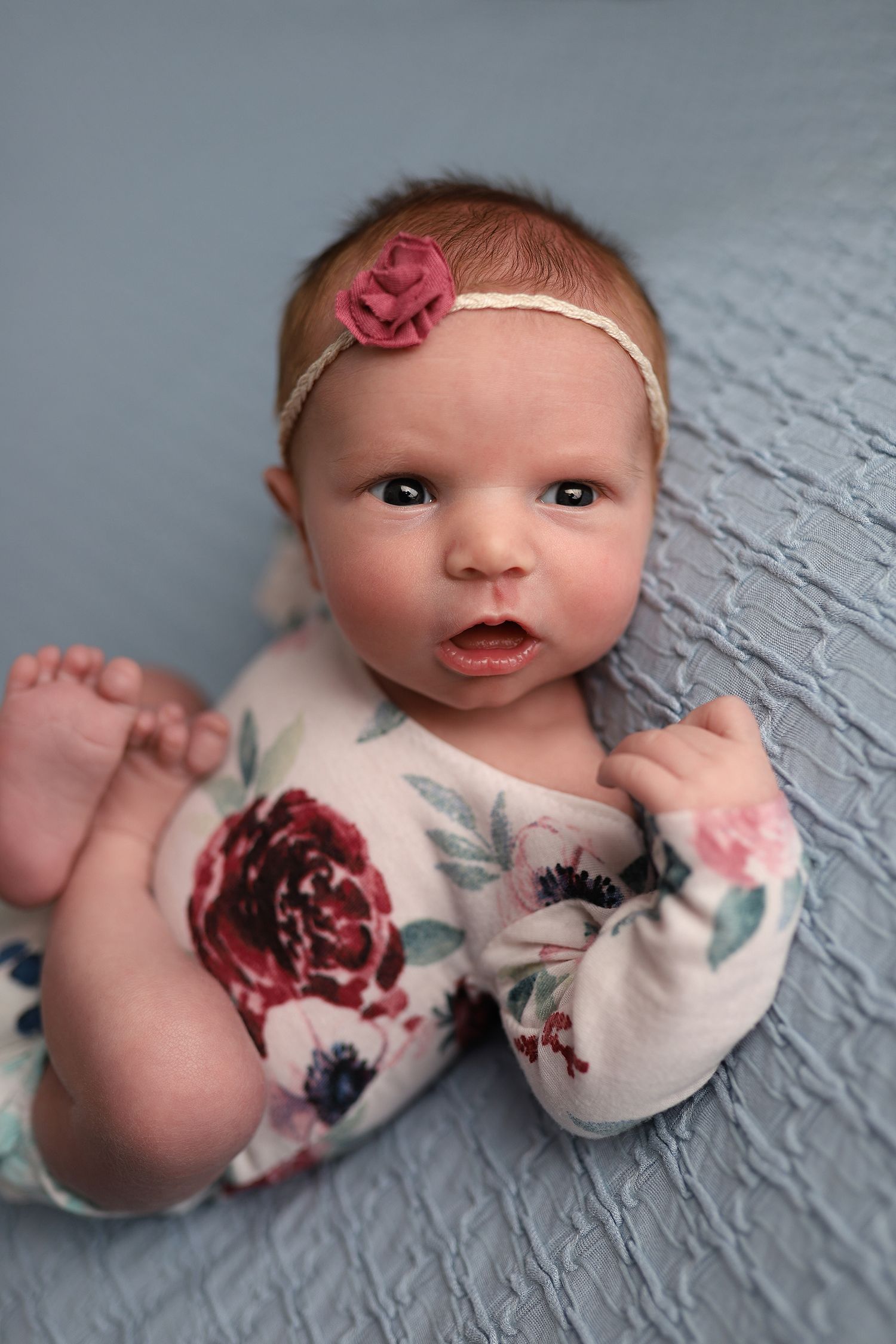 Newborn baby girl in floral romper and pink rose headband lying on blue blanket, looking at camera.