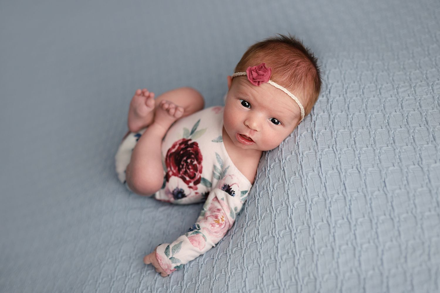 Newborn baby girl in floral outfit and pink headband lying on blue textured blanket during professional photo session. Orem newborn photos