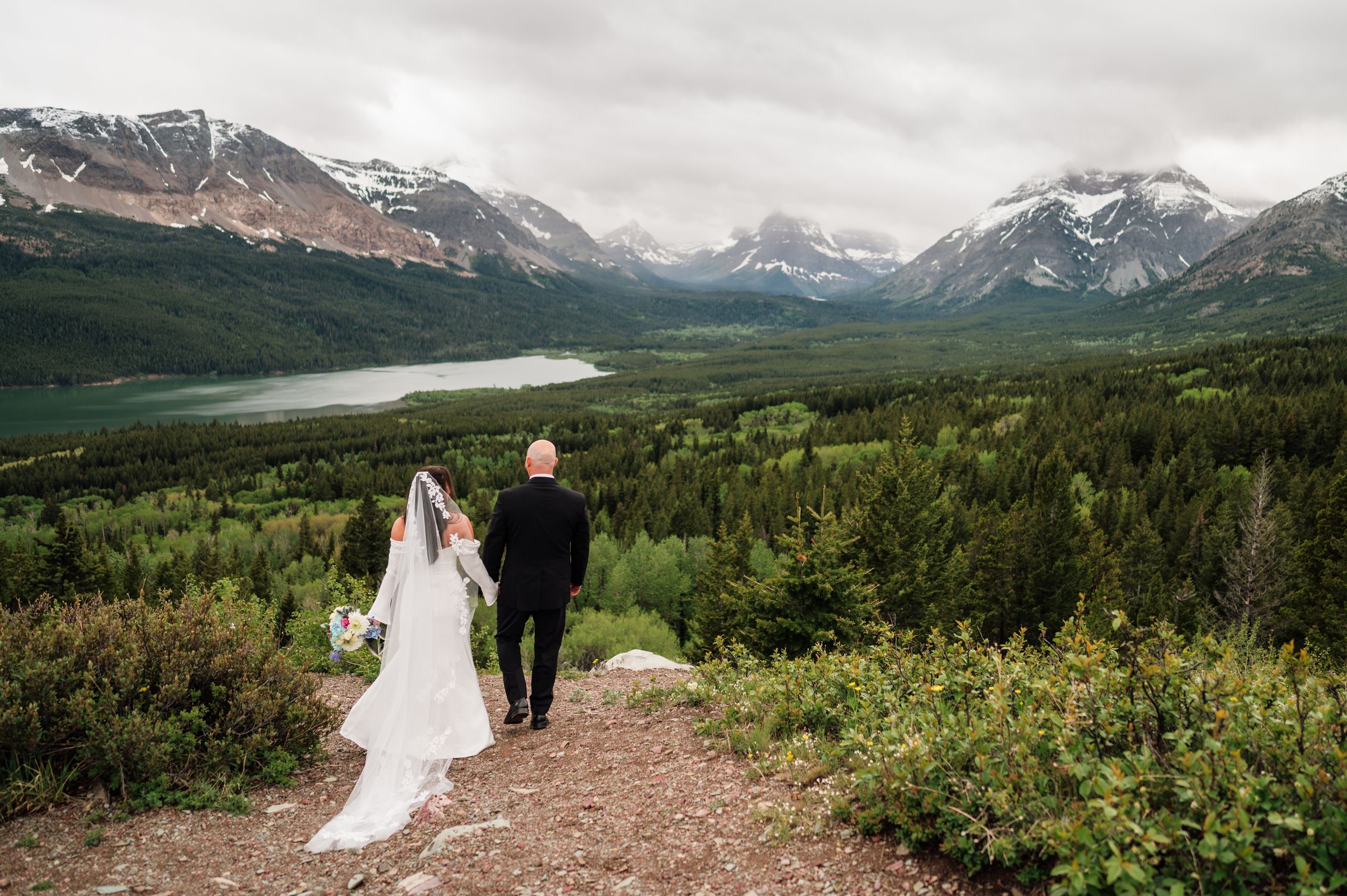 Bride and groom holding hands overlooking a stunning mountain valley with a lake and snow-capped peaks in the background.