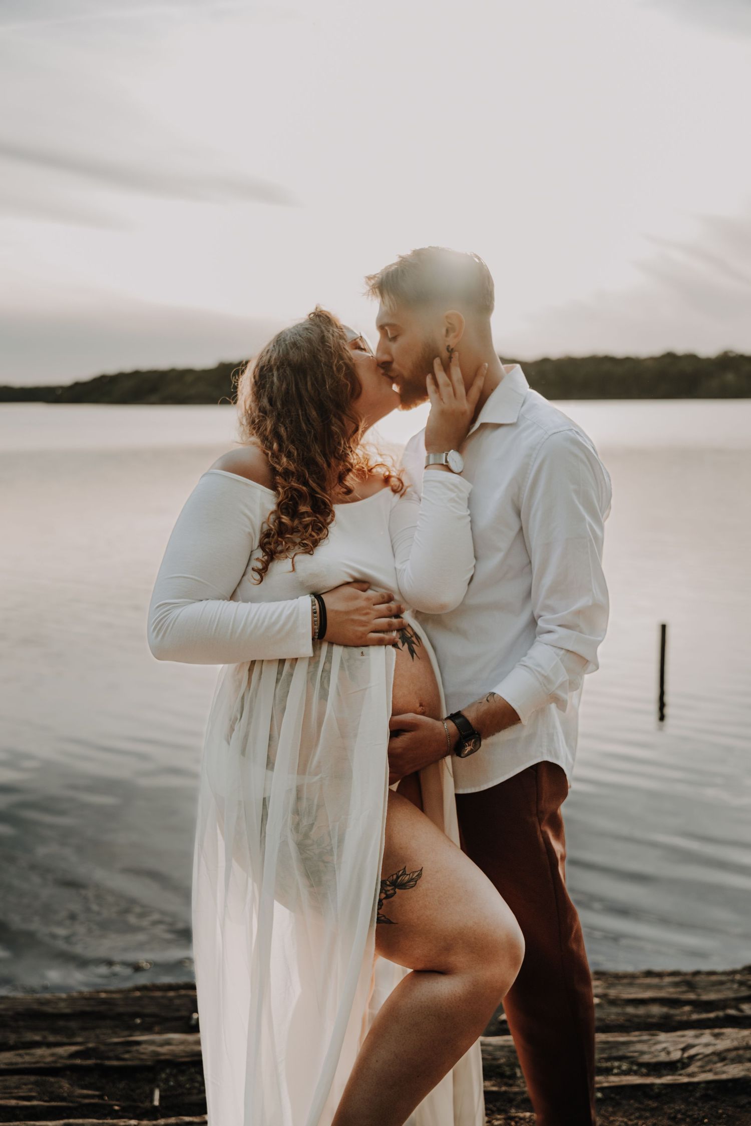 Couple sharing a romantic kiss on a lakeside dock at sunset, woman in flowing white maternity dress.