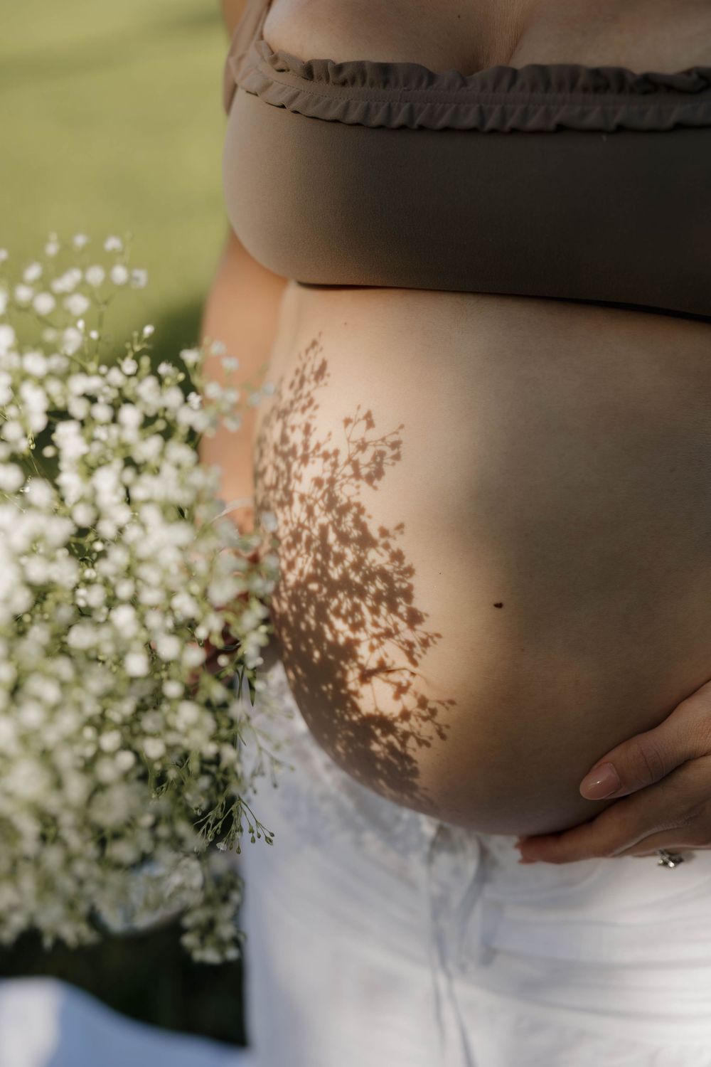 Pregnant woman holding baby bump with floral shadow pattern from white baby's breath flowers in soft natural light.