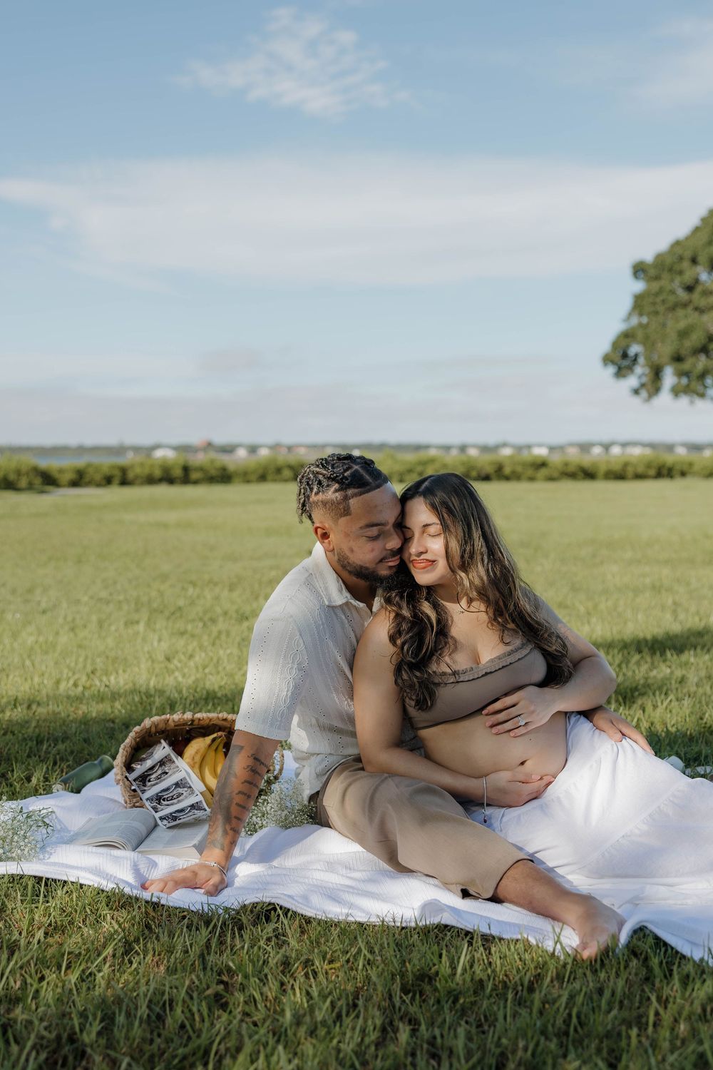 Couple sitting on a blanket in a green field, man kissing pregnant woman's cheek while cradling her baby bump.