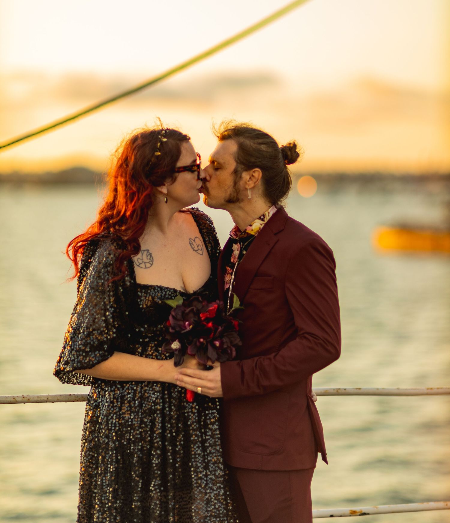 A couple shares a romantic kiss at sunset on a waterfront dock, she in a black dress, he in a burgundy suit.