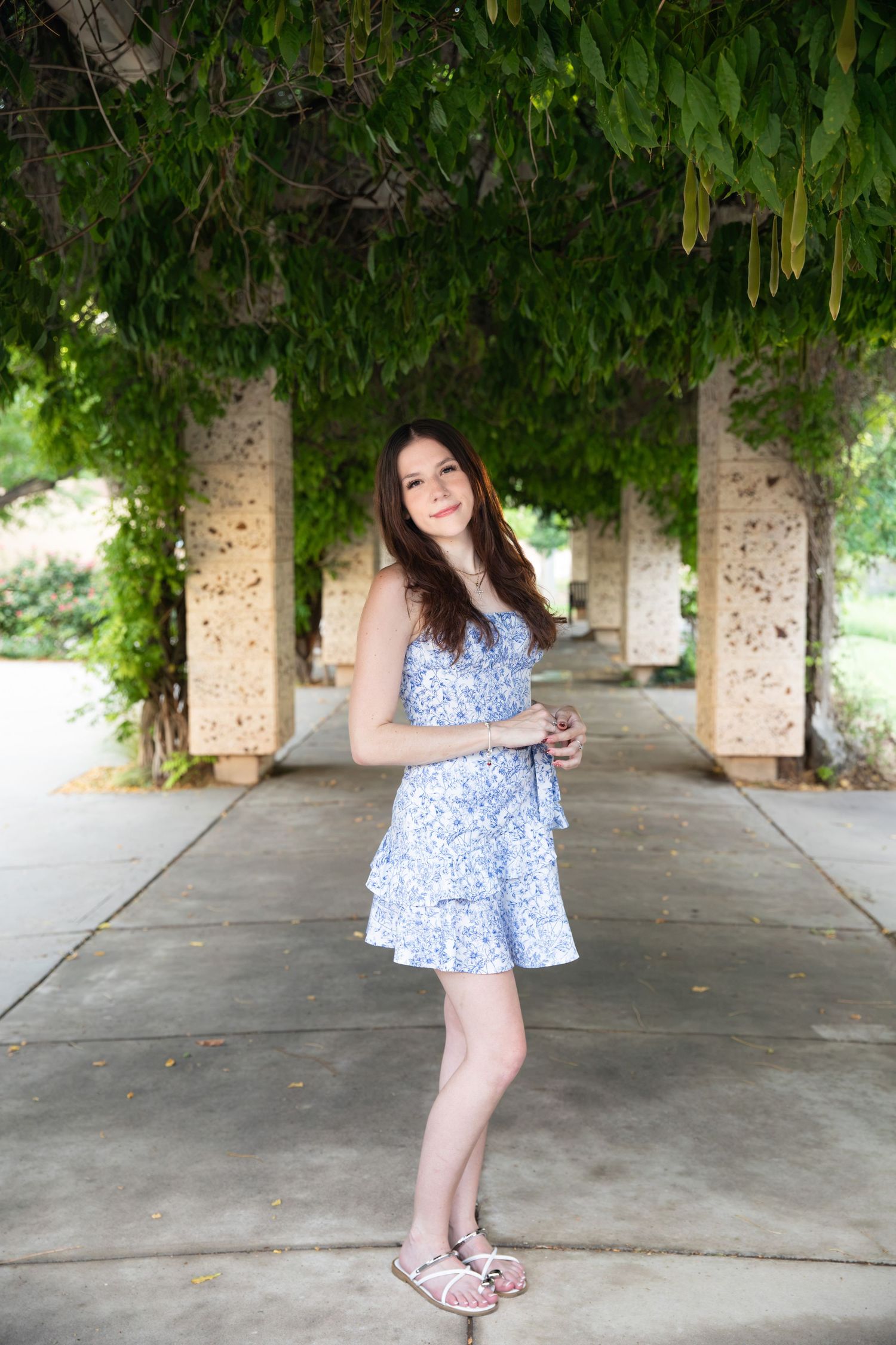 Young woman in blue floral dress posing under vine-covered pergola columns outdoors.