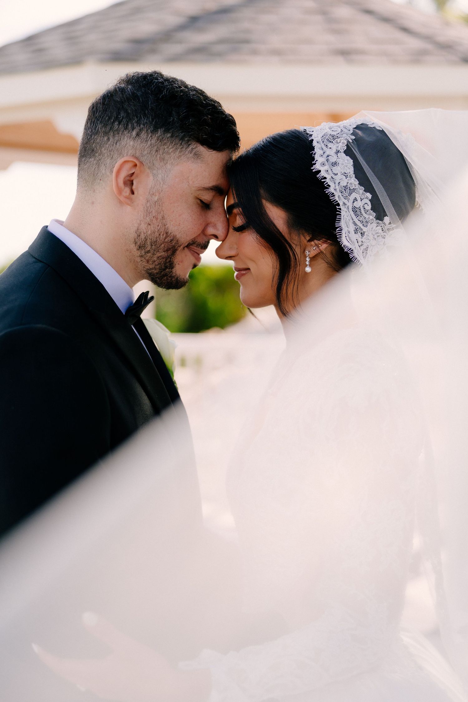 Bride and groom share a romantic moment in a white gazebo, bride wearing lace gown, groom in black tuxedo.