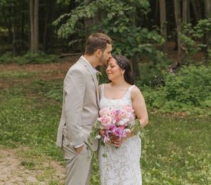 Bride and groom share a tender moment outdoors in a forest, bride holding a pink bouquet in a lace gown.