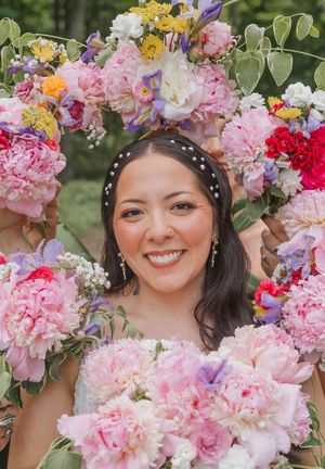 Smiling bride framed by colorful floral arch with pink peonies, wearing pearl headband at outdoor wedding.