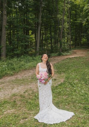 Laughing bride in lace wedding gown holding pink bouquet stands on forest path surrounded by lush green trees.