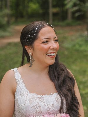 Smiling bride in white lace dress with pearl hair accessories holding pink bouquet outdoors in wooded setting.