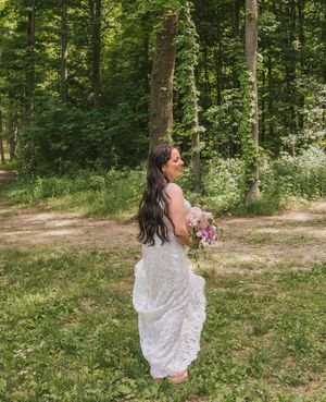 Bride in white lace gown holding pink bouquet, smiling in a lush green forest setting.