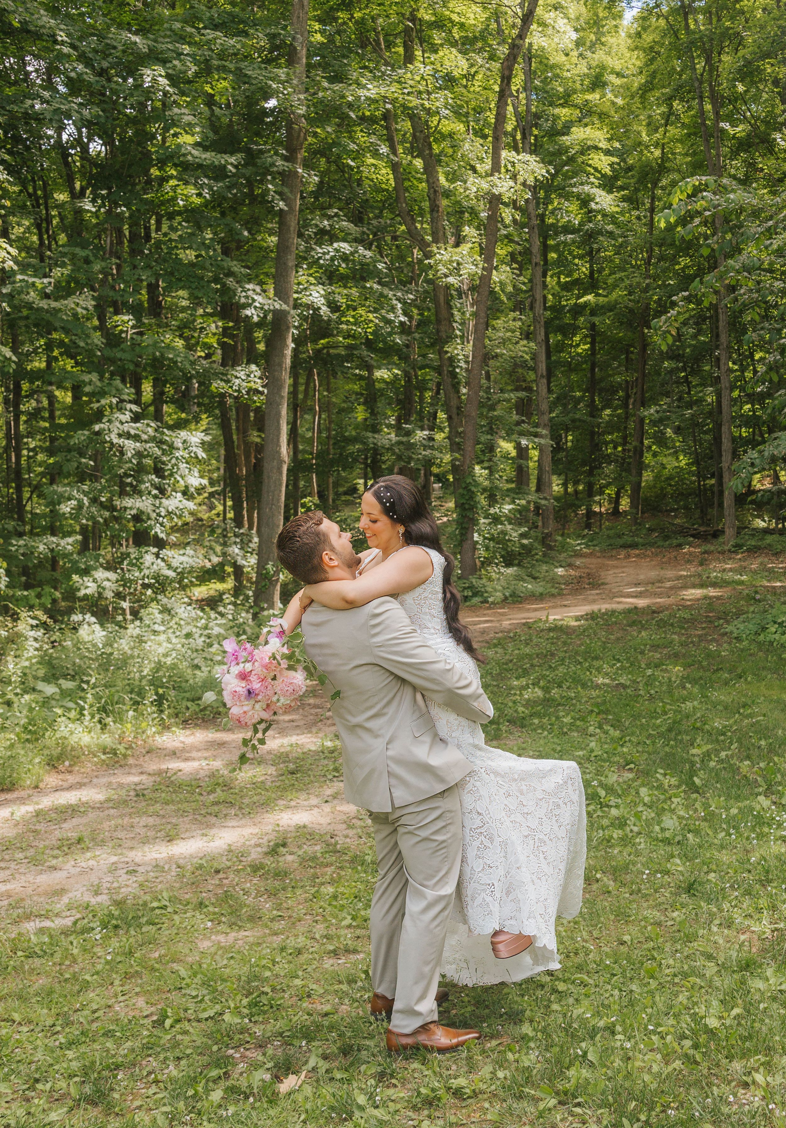 Groom lifts bride in white lace dress holding pink flowers, sharing a romantic moment in a lush green forest clearing.